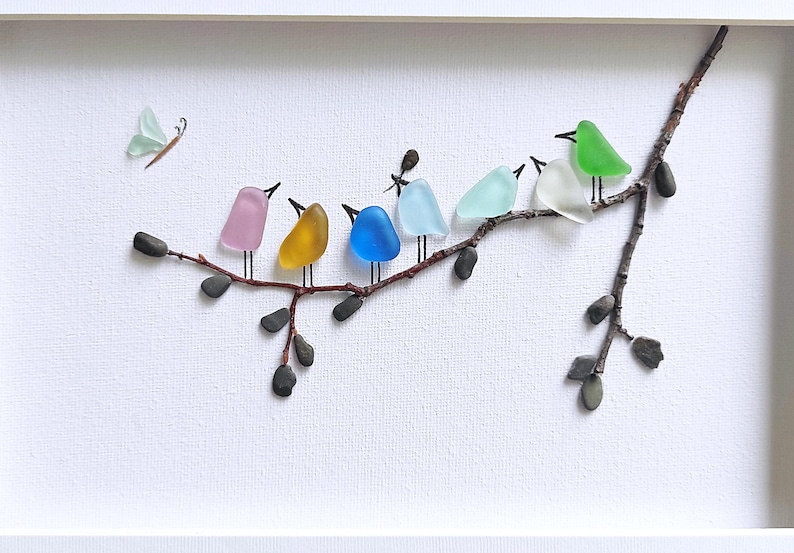 Op de afbeelding: Een ingelijst kunstwerk met een tak waarop kleurrijke glazen vogels zitten. De vogels zijn in roze, geel, blauw, lichtblauw, groen en wit. Er is ook een glazen vlinder aanwezig. Het kunstwerk staat tegen een witte achtergrond.