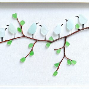 May include: A framed artwork featuring a branch with glass birds and green glass leaves. The birds are light blue, and the leaves are a vibrant green. The branch is brown, and the background is white.