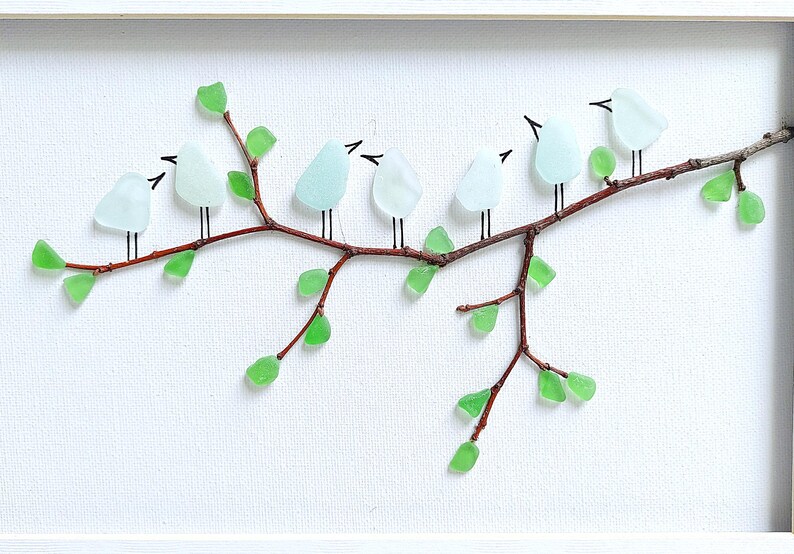 May include: A framed artwork featuring a branch with glass birds and green glass leaves. The birds are light blue, and the leaves are a vibrant green. The branch is brown, and the background is white.