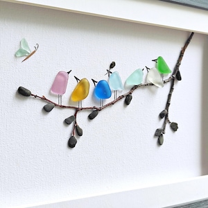 Op de afbeelding: Een ingelijst kunstwerk met een tak met kleurrijke glazen vogels en een vlinder. De vogels zijn in tinten roze, geel, blauw, lichtblauw, groen en wit. Het kunstwerk staat tegen een witte achtergrond en is ingelijst in een grijze lijst.