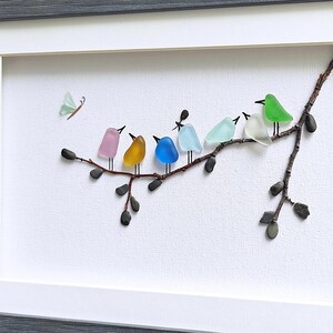 Op de afbeelding: Een ingelijst kunstwerk met een tak met kleurrijke glazen vogels en een vlinder. De vogels zijn in roze, geel, blauw, lichtblauw, wit en groen. Het kunstwerk staat tegen een witte achtergrond.