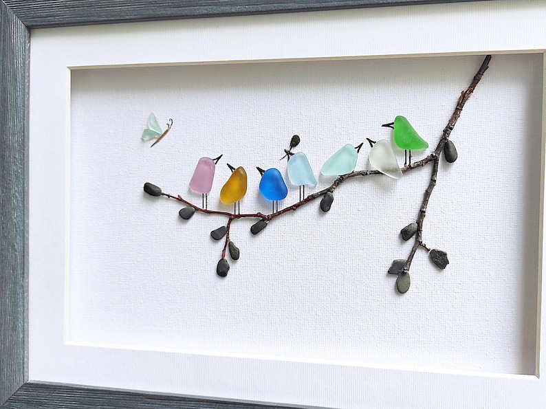 Op de afbeelding: Een ingelijst kunstwerk met een tak met kleurrijke glazen vogels en een vlinder. De vogels zijn in roze, geel, blauw, lichtblauw, wit en groen. Het kunstwerk staat tegen een witte achtergrond.