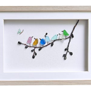 Op de afbeelding: Ingelijst kunstwerk met een tak met kleurrijke glazen vogels. De vogels zijn roze, geel, blauw, lichtblauw, groen en wit. Er is ook een lichtgroene vlinder aanwezig. Het kunstwerk staat tegen een witte achtergrond en is ingelijst in een licht houten frame.