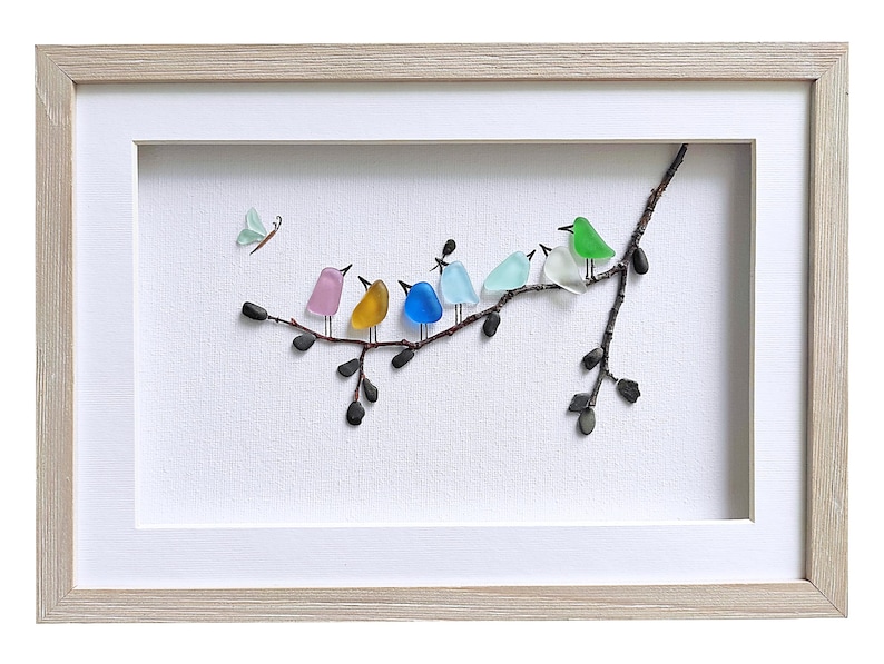 Op de afbeelding: Ingelijst kunstwerk met een tak met kleurrijke glazen vogels. De vogels zijn roze, geel, blauw, lichtblauw, groen en wit. Er is ook een lichtgroene vlinder aanwezig. Het kunstwerk staat tegen een witte achtergrond en is ingelijst in een licht houten frame.