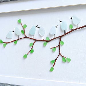 May include: A framed artwork featuring a branch with seven light blue glass birds perched on it. The branch is brown, with green glass leaves. The birds have black beaks and legs. The background is white, and the frame is gray.