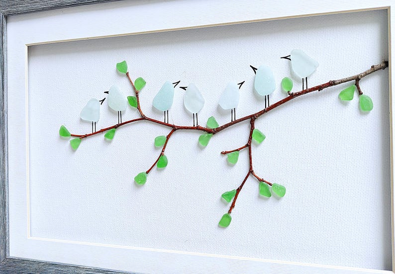 May include: A framed artwork featuring a branch with seven light blue glass birds perched on it. The branch is brown, with green glass leaves. The birds have black beaks and legs. The background is white, and the frame is gray.