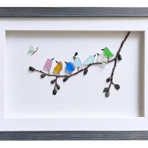 Op de afbeelding: Een ingelijst kunstwerk met een tak met zeven kleurrijke vogels gemaakt van zee glas. De vogels zijn roze, geel, blauw, lichtblauw, wit en groen. Een kleine vlinder staat in de linkerbovenhoek. De lijst is grijs.