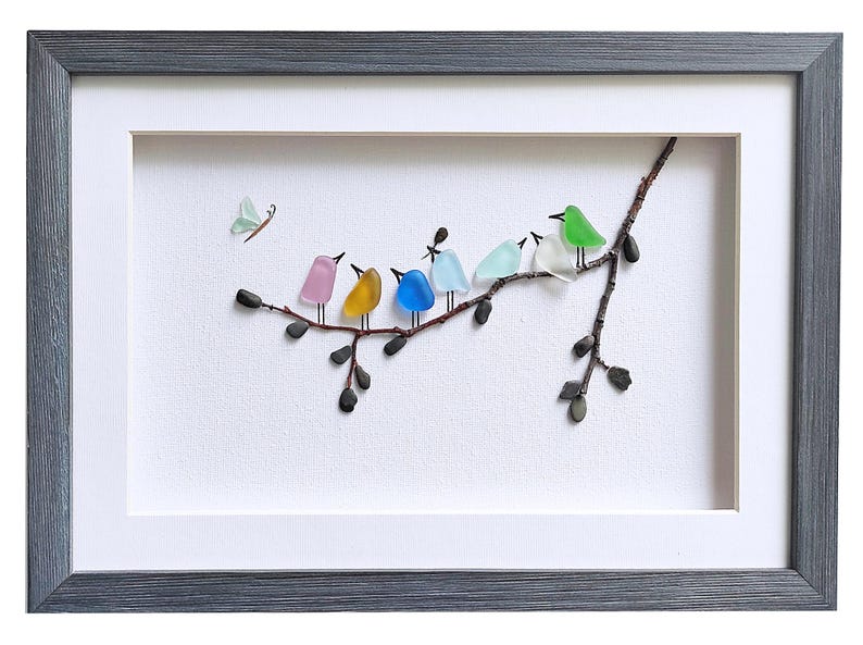 Op de afbeelding: Een ingelijst kunstwerk met een tak met zeven kleurrijke vogels gemaakt van zee glas. De vogels zijn roze, geel, blauw, lichtblauw, wit en groen. Een kleine vlinder staat in de linkerbovenhoek. De lijst is grijs.