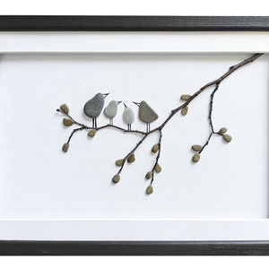 May include: A framed artwork featuring a branch with small leaves and four birds made of stones. The birds are gray, white, and brown. The background is white.