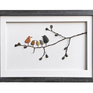 Op de afbeelding: Een ingelijst kunstwerk van kiezelsteen dat een familie vogels op een tak toont. De vogels zijn gemaakt van verschillende gekleurde stenen, waaronder oranje, bruin, grijs en zwart. De tak is gemaakt van een natuurlijke twijg.