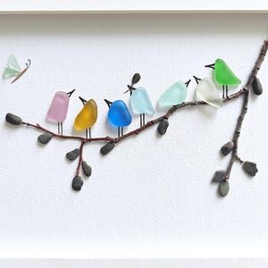 Op de afbeelding: Een ingelijst kunstwerk met kleurrijke glazen vogels die op een tak zitten. De vogels zijn in verschillende kleuren, waaronder roze, geel, blauw en groen. Er is ook een glazen vlinder aanwezig. Het kunstwerk staat tegen een witte achtergrond.