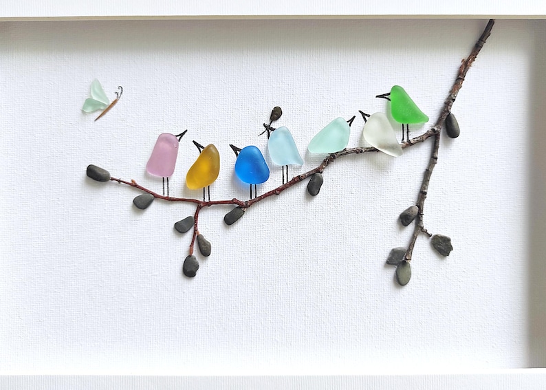Op de afbeelding: Een ingelijst kunstwerk met kleurrijke glazen vogels die op een tak zitten. De vogels zijn in verschillende kleuren, waaronder roze, geel, blauw en groen. Er is ook een glazen vlinder aanwezig. Het kunstwerk staat tegen een witte achtergrond.