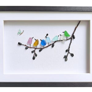 Op de afbeelding: Ingelijst kunstwerk met een tak waarop kleurrijke glazen vogels zitten. De vogels zijn in verschillende kleuren, waaronder roze, geel, blauw en groen. Er is ook een kleine vlinder aanwezig. Het kunstwerk is ingelijst in een zwarte lijst met een witte passe-partout.