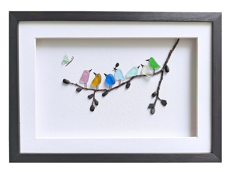 Op de afbeelding: Ingelijst kunstwerk met een tak waarop kleurrijke glazen vogels zitten. De vogels zijn in verschillende kleuren, waaronder roze, geel, blauw en groen. Er is ook een kleine vlinder aanwezig. Het kunstwerk is ingelijst in een zwarte lijst met een witte passe-partout.