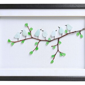 May include: Framed artwork featuring a branch with eight light blue glass birds perched on it. The branch has green glass leaves. The artwork is set against a white background and framed in a black frame.