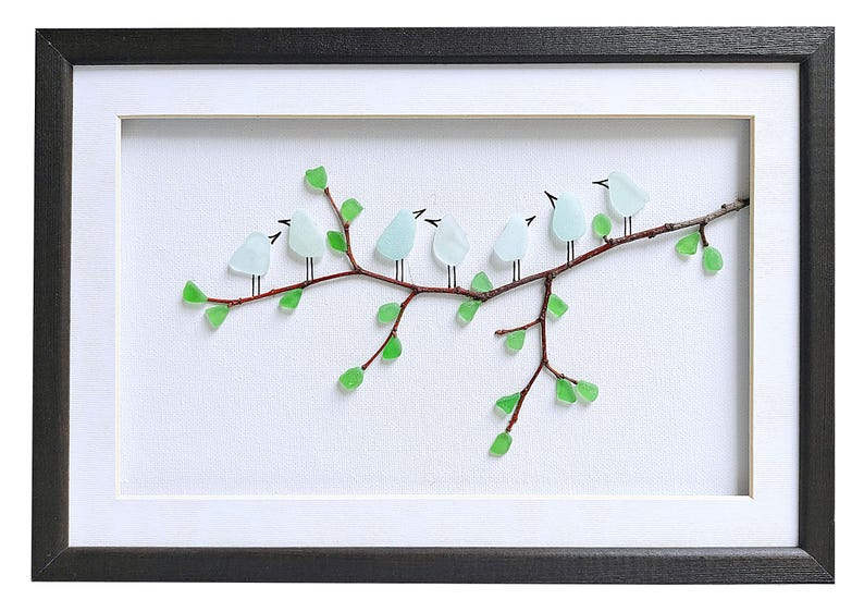 May include: Framed artwork featuring a branch with eight light blue glass birds perched on it. The branch has green glass leaves. The artwork is set against a white background and framed in a black frame.