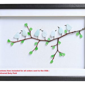 May include: Framed artwork featuring a branch with seven light blue glass birds perched on it. The branch is brown with green glass leaves. The artwork is set against a white background, framed in black and white.