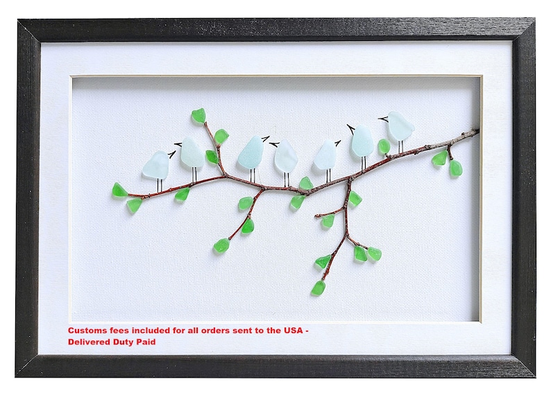 May include: Framed artwork featuring a branch with seven light blue glass birds perched on it. The branch is brown with green glass leaves. The artwork is set against a white background, framed in black and white.