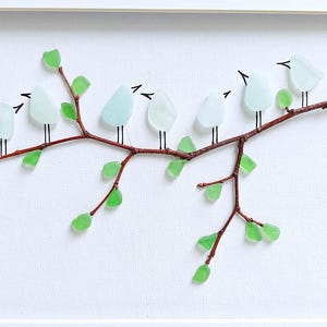 May include: A framed artwork featuring a branch with seven light blue glass birds perched on it. The branch is brown with green glass leaves. The background is white, and the frame is white.