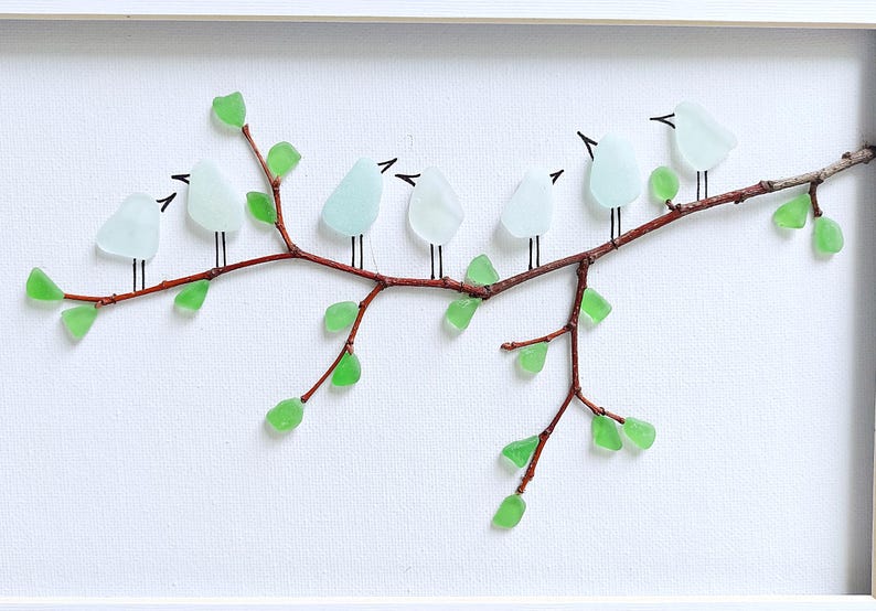 May include: A framed artwork featuring a branch with seven light blue glass birds perched on it. The branch is brown with green glass leaves. The background is white, and the frame is white.
