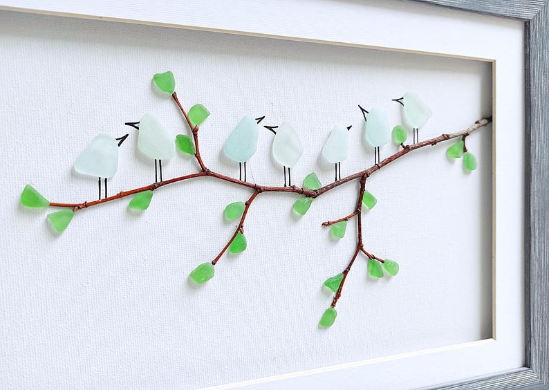 May include: A framed artwork featuring a branch with light blue glass birds perched on it. The branch has green glass leaves. The artwork is set against a white background and framed in a gray wooden frame.