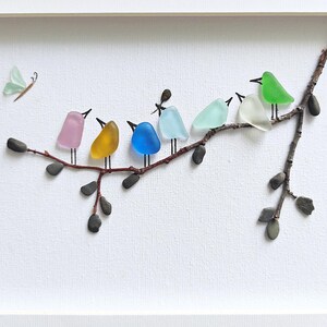 Op de afbeelding: Een ingelijst kunstwerk met een tak waarop zeven kleurrijke glazen vogels zitten. De vogels zijn in verschillende kleuren, waaronder roze, geel, blauw en groen. Er is ook een kleine vlinder aanwezig, wat een speels tintje geeft.