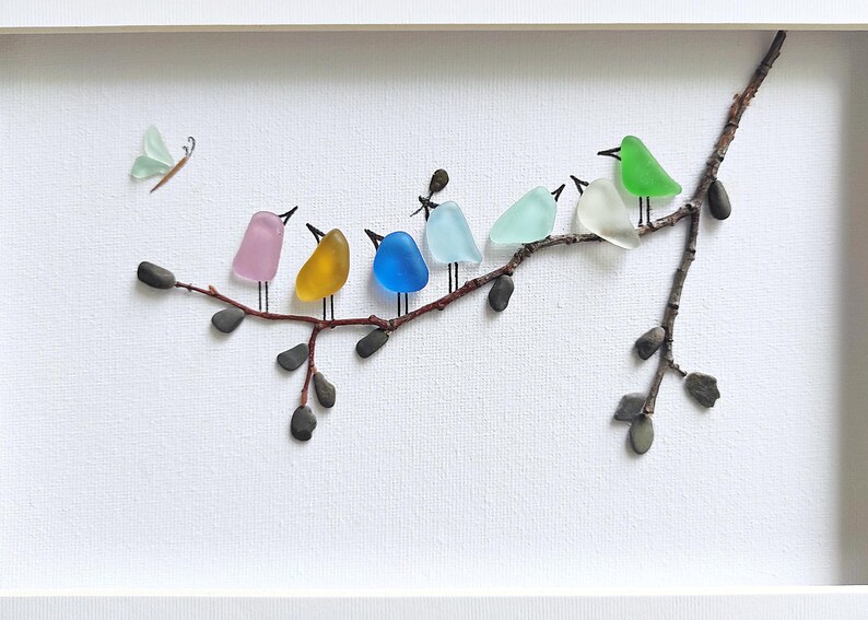 Op de afbeelding: Een ingelijst kunstwerk met een tak waarop zeven kleurrijke glazen vogels zitten. De vogels zijn in verschillende kleuren, waaronder roze, geel, blauw en groen. Er is ook een kleine vlinder aanwezig, wat een speels tintje geeft.