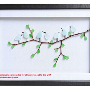 May include: Framed artwork featuring a branch with seven light blue glass birds perched on it. The branch is brown with green glass leaves. The artwork is set against a white background, framed in black and white.