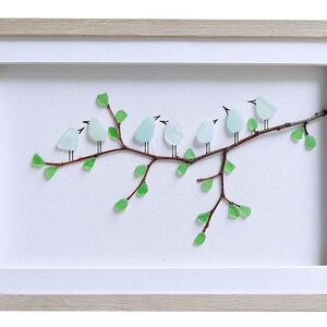 May include: A framed artwork featuring a branch with seven light blue glass birds perched on it. The branch has green glass leaves. The artwork is set against a white background and framed in a light wood frame.