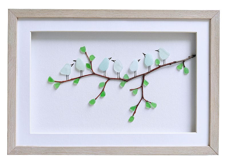 May include: A framed artwork featuring a branch with seven light blue glass birds perched on it. The branch has green glass leaves. The artwork is set against a white background and framed in a light wood frame.