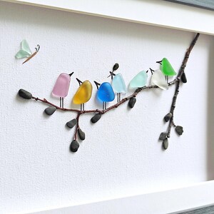 Op de afbeelding: Een ingelijst kunstwerk met kleurrijke glazen vogels die op een tak zitten. De vogels hebben verschillende kleuren, waaronder roze, geel, blauw en groen. Er is ook een glazen vlinder aanwezig. Het kunstwerk staat tegen een witte achtergrond.