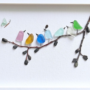 Op de afbeelding: Een ingelijst kunstwerk met een tak waarop zeven kleurrijke glazen vogels zitten. Een lichtgroene glazen vlinder vliegt in de buurt. De vogels zijn in verschillende kleuren, waaronder roze, geel, blauw en groen, tegen een witte achtergrond.