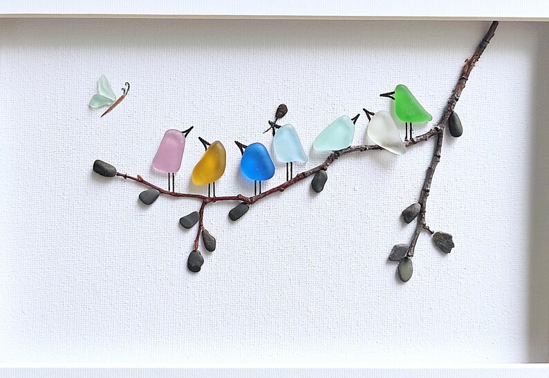 Op de afbeelding: Een ingelijst kunstwerk met een tak waarop zeven kleurrijke glazen vogels zitten. Een lichtgroene glazen vlinder vliegt in de buurt. De vogels zijn in verschillende kleuren, waaronder roze, geel, blauw en groen, tegen een witte achtergrond.
