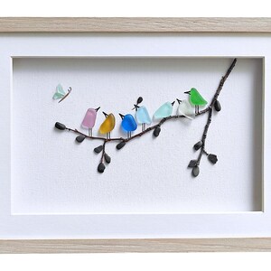Op de afbeelding: Ingelijst kunstwerk met een tak met kleurrijke glazen vogels. De vogels zijn in tinten roze, geel, blauw, lichtblauw, groen en wit. Een lichtgroene vlinder bevindt zich in de linkerbovenhoek. Het kunstwerk zit in een witte passe-partout met een licht houten lijst.