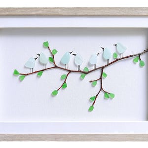 May include: A framed artwork featuring a branch with seven light blue bird figures made of glass. The branch is brown with green glass leaves. The artwork is set against a white background and framed in a light wood frame.