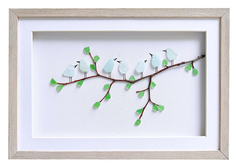 May include: A framed artwork featuring a branch with seven light blue bird figures made of glass. The branch is brown with green glass leaves. The artwork is set against a white background and framed in a light wood frame.