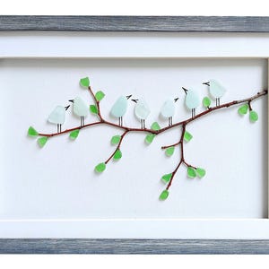 May include: Framed artwork featuring a branch with light blue glass birds perched on it. The branch has green glass leaves. The artwork is set against a white background and framed in a gray wooden frame.