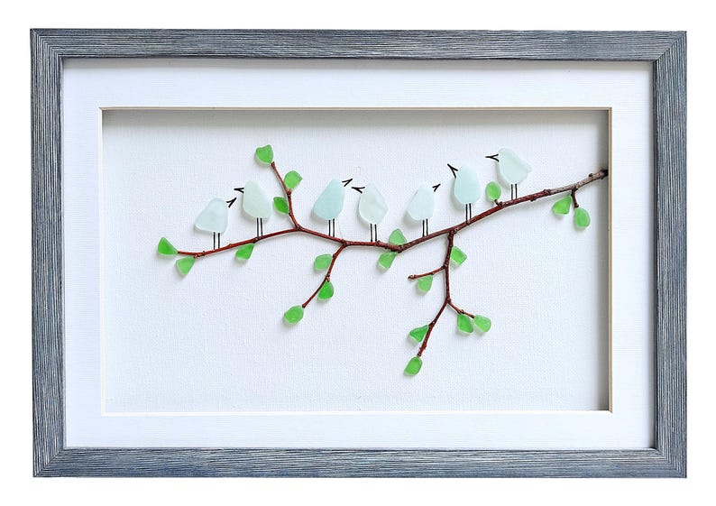 May include: Framed artwork featuring a branch with light blue glass birds perched on it. The branch has green glass leaves. The artwork is set against a white background and framed in a gray wooden frame.