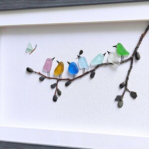Op de afbeelding: Een ingelijst kunstwerk met een tak met kleurrijke glazen vogels en een vlinder. De vogels zijn in roze, geel, blauw en groen. Het kunstwerk staat tegen een witte achtergrond en is grijs omlijst.