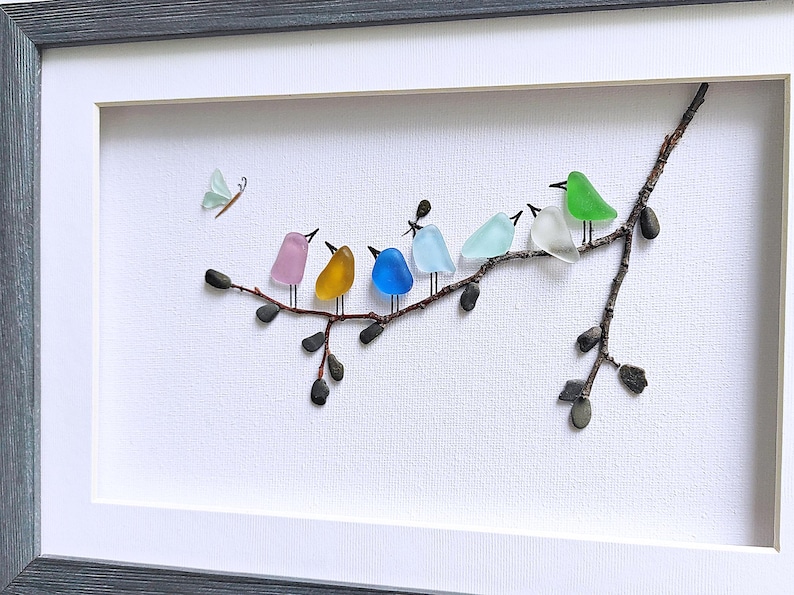 Op de afbeelding: Een ingelijst kunstwerk met een tak met kleurrijke glazen vogels en een vlinder. De vogels zijn in roze, geel, blauw en groen. Het kunstwerk staat tegen een witte achtergrond en is grijs omlijst.