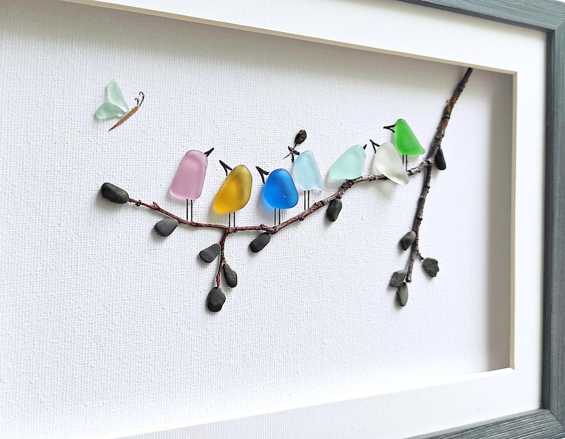 Op de afbeelding: Een ingelijst kunstwerk met kleurrijke glazen vogels op een tak. De vogels zijn in verschillende kleuren, waaronder roze, geel, blauw en groen. Er is ook een vlinder van glas aanwezig. Het kunstwerk staat tegen een witte achtergrond.