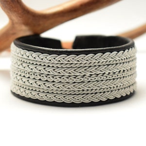 Könnte beinhalten: Ein schwarzes Lederarmband mit einem geflochtenen Silberdesign. Das Armband ist breit und hat einen schlichten, eleganten Look.