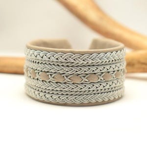 Könnte beinhalten: Ein beigefarbenes Lederarmband mit einem geflochtenen Silberdesign. Das Armband hat eine Reihe von beigefarbenen Lederakzenten.
