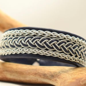 Könnte beinhalten: Ein dunkelblaues Lederarmband mit aufwendigen silbernen geflochtenen Details. Das Armband hat ein gewebtes Muster um das Band, wodurch ein strukturiertes und stilvolles Accessoire entsteht. Das Design ist eine Nahaufnahme, die die Handwerkskunst hervorhebt.