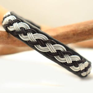 Könnte beinhalten: Nahaufnahme eines schwarzen Lederarmbands mit geflochtenem Design. Das Armband weist ein Muster aus miteinander verwobenen silbernen und dunkelbraunen Strängen auf. Das Armband ist ein stilvolles Accessoire.