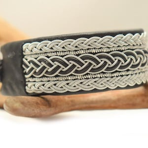 Könnte beinhalten: Ein schwarzes Lederarmband mit aufwendigen, silberfarbenen, geflochtenen Details. Das Armband hat ein zentrales Webmuster und wird mit einem silberfarbenen Verschluss befestigt. Das Armband ist etwa 3,8 cm breit.