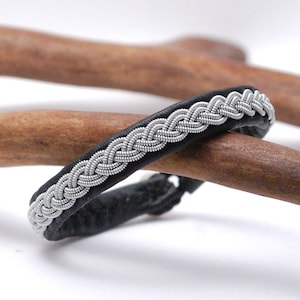 Puede incluir: Un brazalete de cuero negro con un diseño trenzado plateado. El brazalete está sobre una superficie de madera.