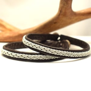 Pode incluir: Duas pulseiras de couro marrom com acabamento trançado prateado. As pulseiras estão empilhadas uma em cima da outra.