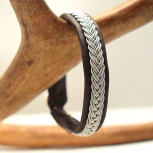 Könnte beinhalten: Dunkelbraunes Lederarmband mit silbernem Zopfmuster. Das Armband wird auf einem hellbraunen Hirschgeweih präsentiert, was den Kontrast zwischen Materialien und Texturen hervorhebt. Nahaufnahme.