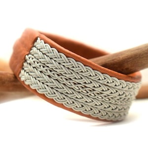 Könnte beinhalten: Ein braunes Lederarmband mit einem geflochtenen Silberdesign. Das Armband liegt auf einer Holzoberfläche.